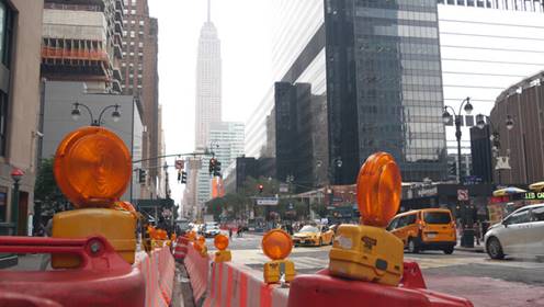 Street view of New York City construction site. The Empire State building rises in the distance, seen between orange safety barriers.